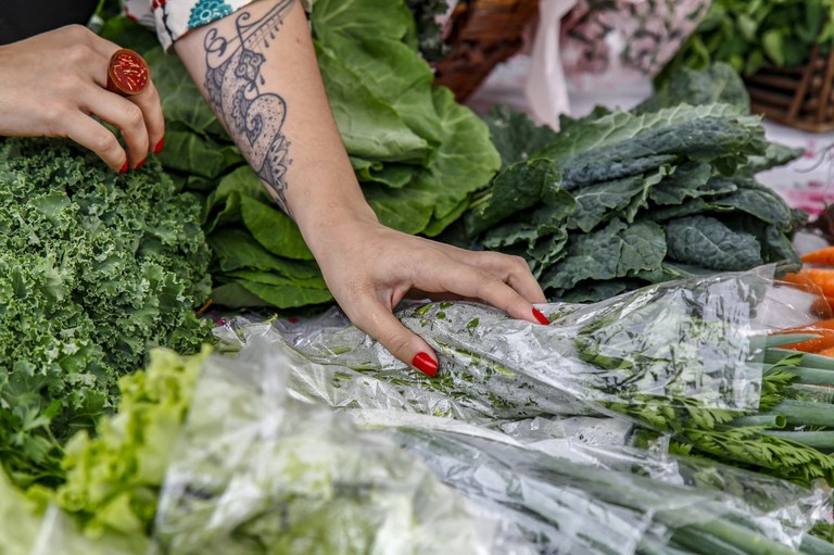 foto mostra mão de pessoa branca com braço tatuado, sob banca de verduras 